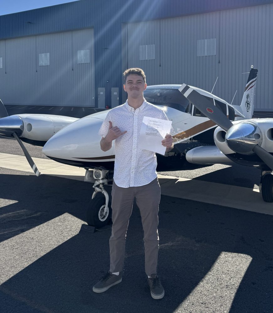 Person standing in front of small airplane outside a hangar on a sunny day.